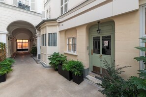 A narrow alleyway with a building on the right, featuring a green door, potted plants, and a small window with barred glass. - Bright Stylish 1 BDR Flat Folies Bergere (Paris)