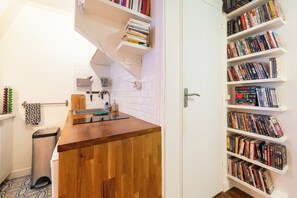 A compact kitchen with a wooden countertop, a sink, and a white tiled backsplash. Adjacent to the kitchen is a white door leading to a room with a bookshelf filled with books.