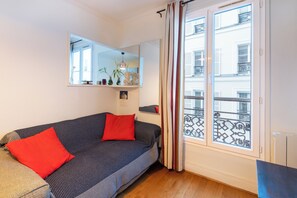 A room with a sofa, two red pillows, a mirror, and a window with a view of buildings. - Bright Stylish 1 BDR Flat Folies Bergere (Paris)