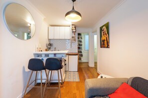 A compact living space with a kitchenette, a dining area with stools, a sofa with cushions, and a hallway leading to another room.