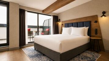 Habitación de hotel con una cama grande, cabecera de madera, una mesita pequeña y vistas a un área exterior con sillas a través de una puerta corredera.