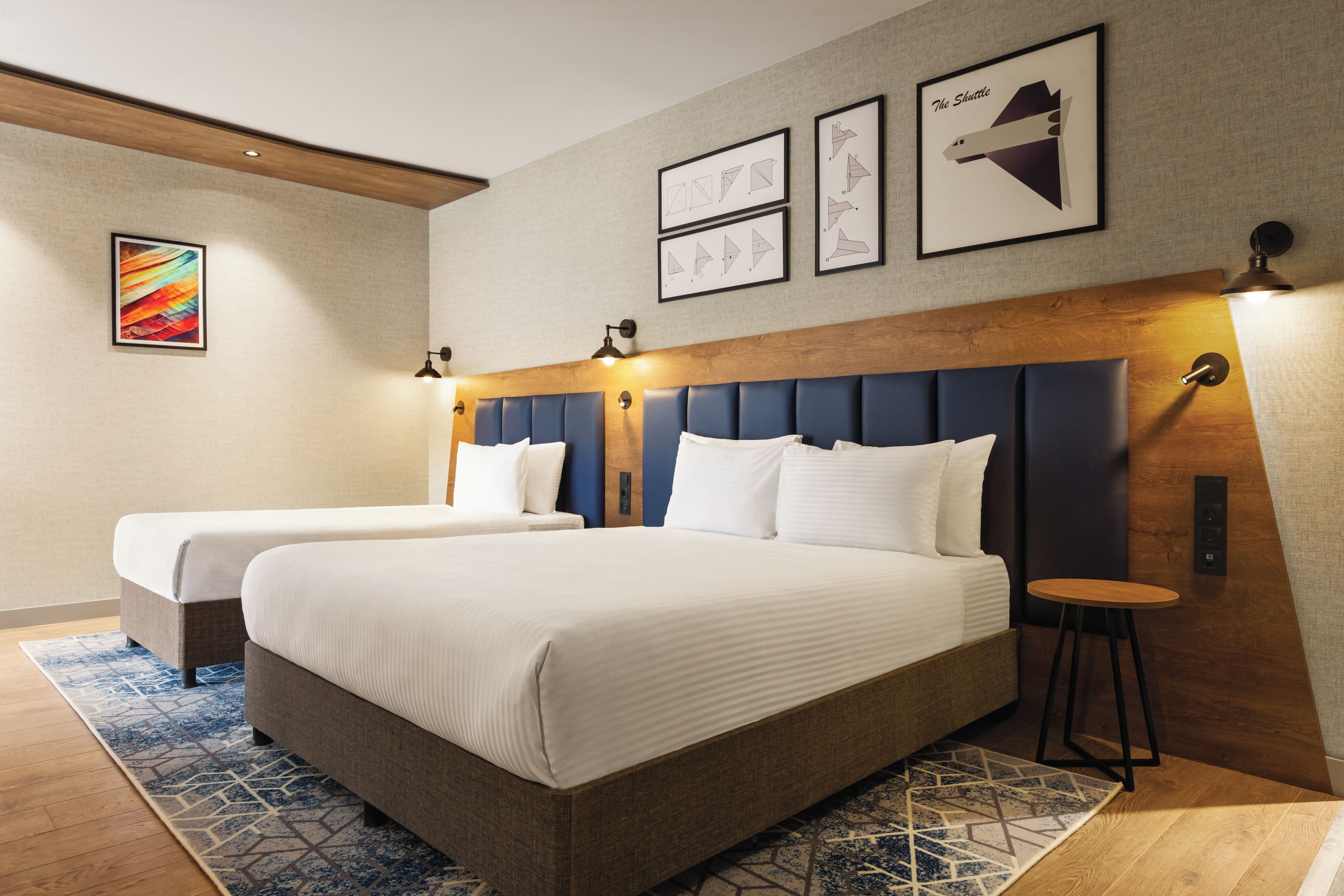 A hotel room with two beds, a wooden headboard, a blue accent wall, and a small round table.