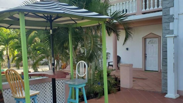 Un patio al aire libre con una mesa redonda y sillas coloridas, un paraguas a rayas y una casa con balcón al fondo.