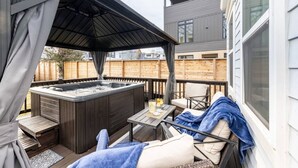 A covered outdoor patio with a hot tub, seating area, and a view of neighboring buildings.
