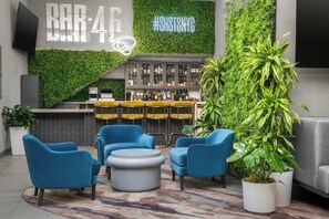 Zona de bar con pared verde, sillas azules, barra y una planta grande en maceta.