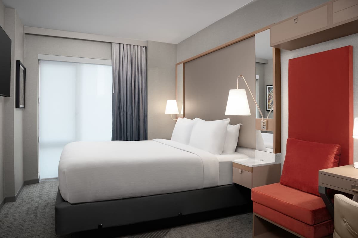 A modern hotel room with a large bed, a desk with a chair, a mirror, and a red accent wall.
