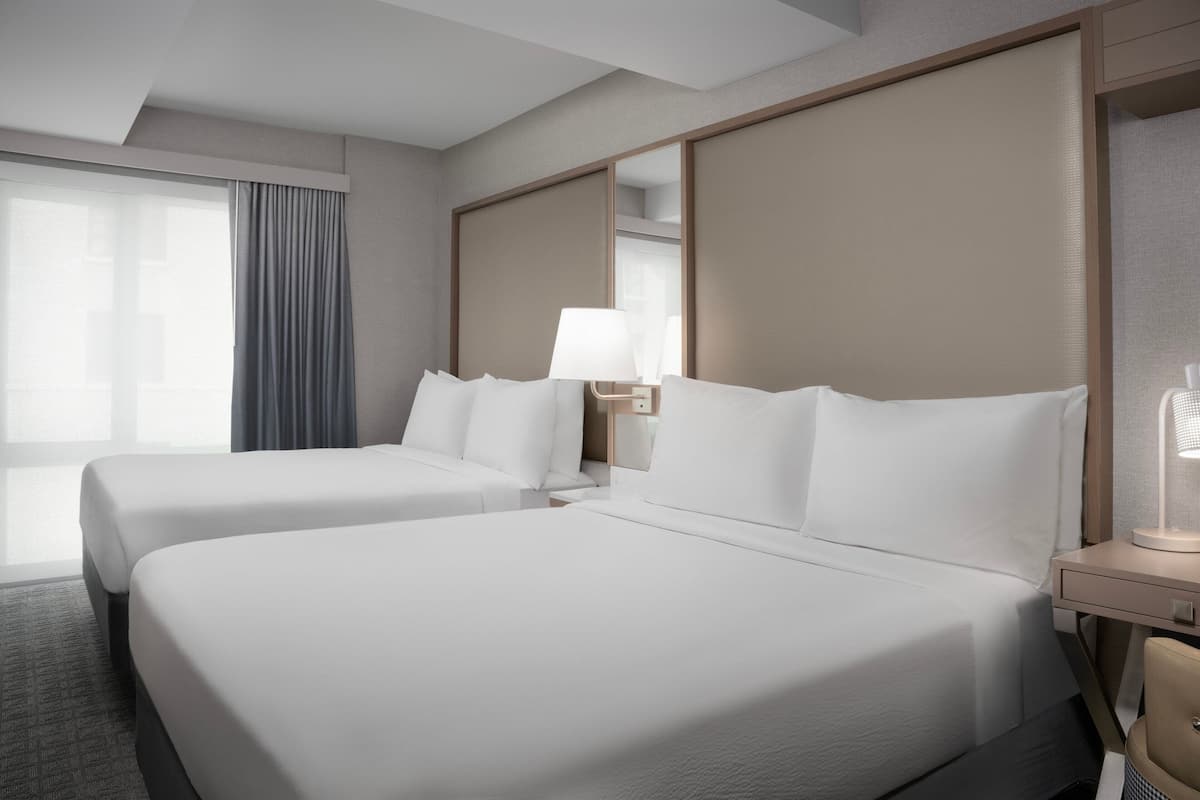 A hotel room with two beds, a large window with curtains, a bedside table with a lamp, and a mirror.