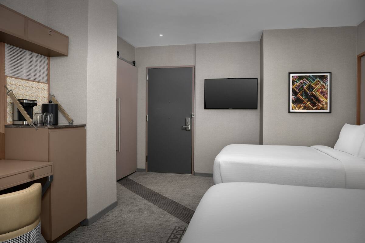 A hotel room with a bed, a desk with a chair, a coffee machine, and a TV mounted on the wall.