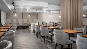 Un restaurant moderne doté d’un sol à motifs hexagonaux, de chaises à revêtement gris, de tables en bois et d’une colonne centrale ornée de lignes verticales.