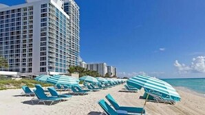 A beach with rows of blue lounge chairs and matching umbrellas, a tall hotel building in the background, and clear blue skies.