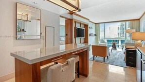 A modern hotel room with a large window, a flat-screen TV, a wooden desk, and a seating area with a chair and a sofa.