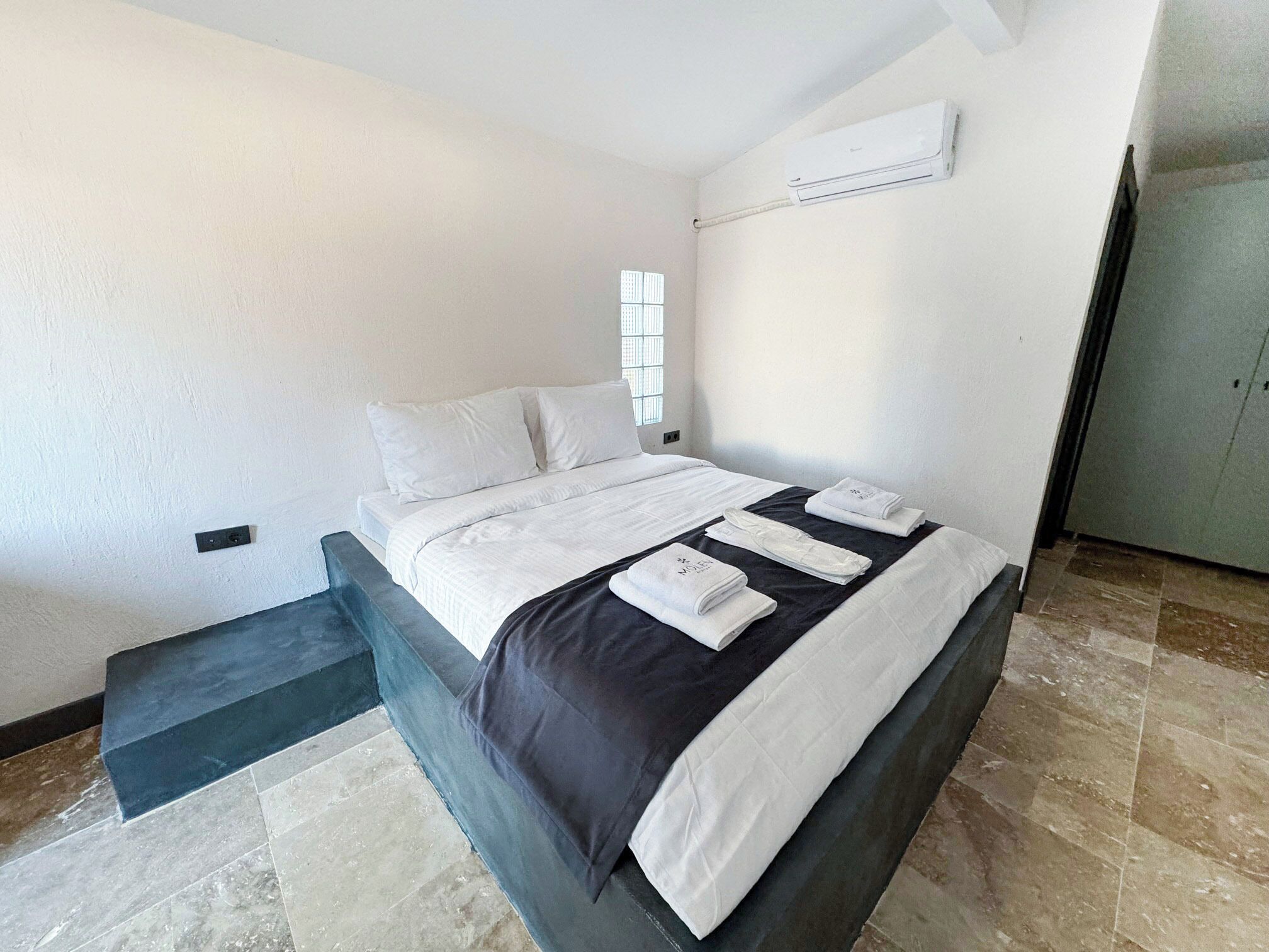 A hotel room with a bed, a bench, an air conditioning unit, and a wardrobe.