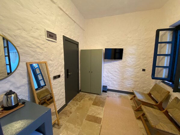 A room with a white textured wall, a grey wardrobe, a television mounted above a door, a small table with a kettle, and a wooden chair.
