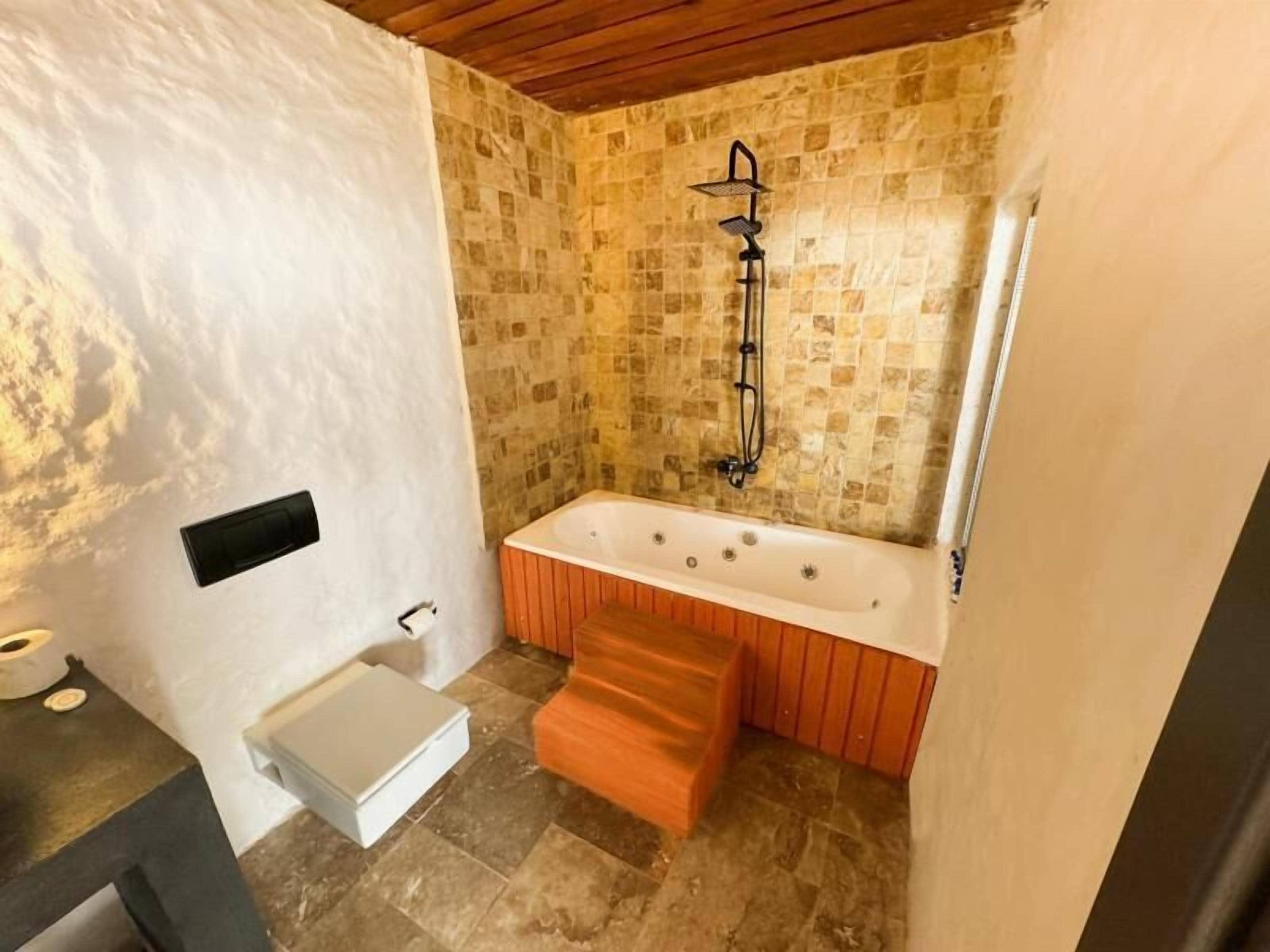 A bathroom with a stone-tiled floor, a wooden bathtub, a shower with a black hose, and a white sink.