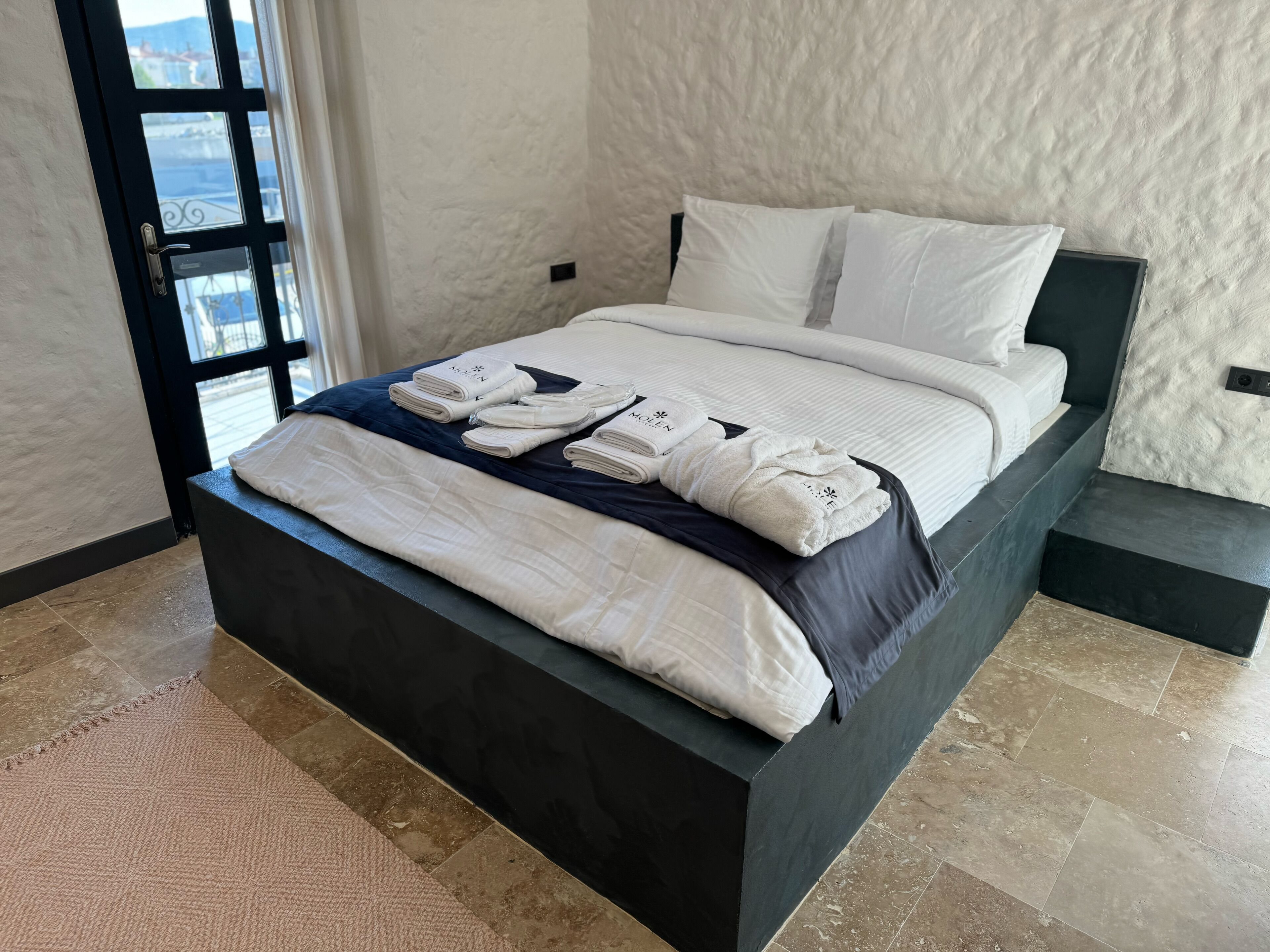 A neatly made bed with white linens and towels, a dark headboard, and a view through a window.