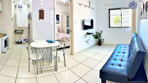 A compact living space with a kitchenette, a dining table with chairs, a sofa, a TV, and a potted plant. - Edf. Gales de Pajuçara - Apt. 105 (Pajuçara Maceió)