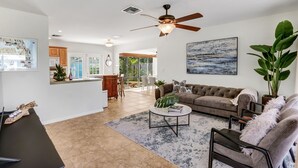A spacious living area with a sofa, coffee table, and a ceiling fan. There is a kitchen in the background with wooden cabinets and a dining area with chairs. - Work & Play Retreat. Private Office. 1GB Internet (Oakland Park)