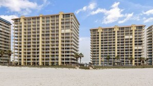 Un hotel frente a la playa con varios pisos, balcones y una playa arenosa al frente.