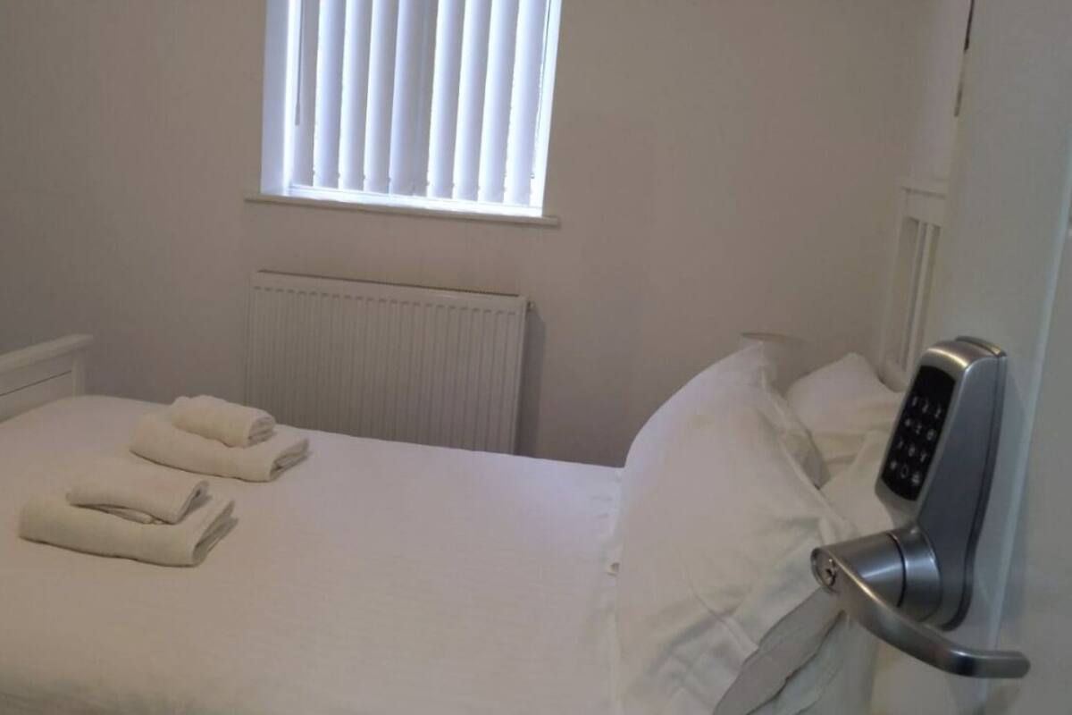 A hotel room with a bed, two rolled towels, a window with blinds, a radiator, and a modern door lock.