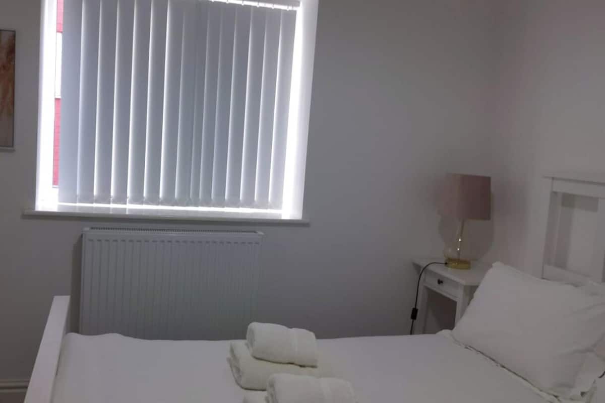 A hotel room with a bed, a nightstand with a lamp, a window with blinds, and a radiator.