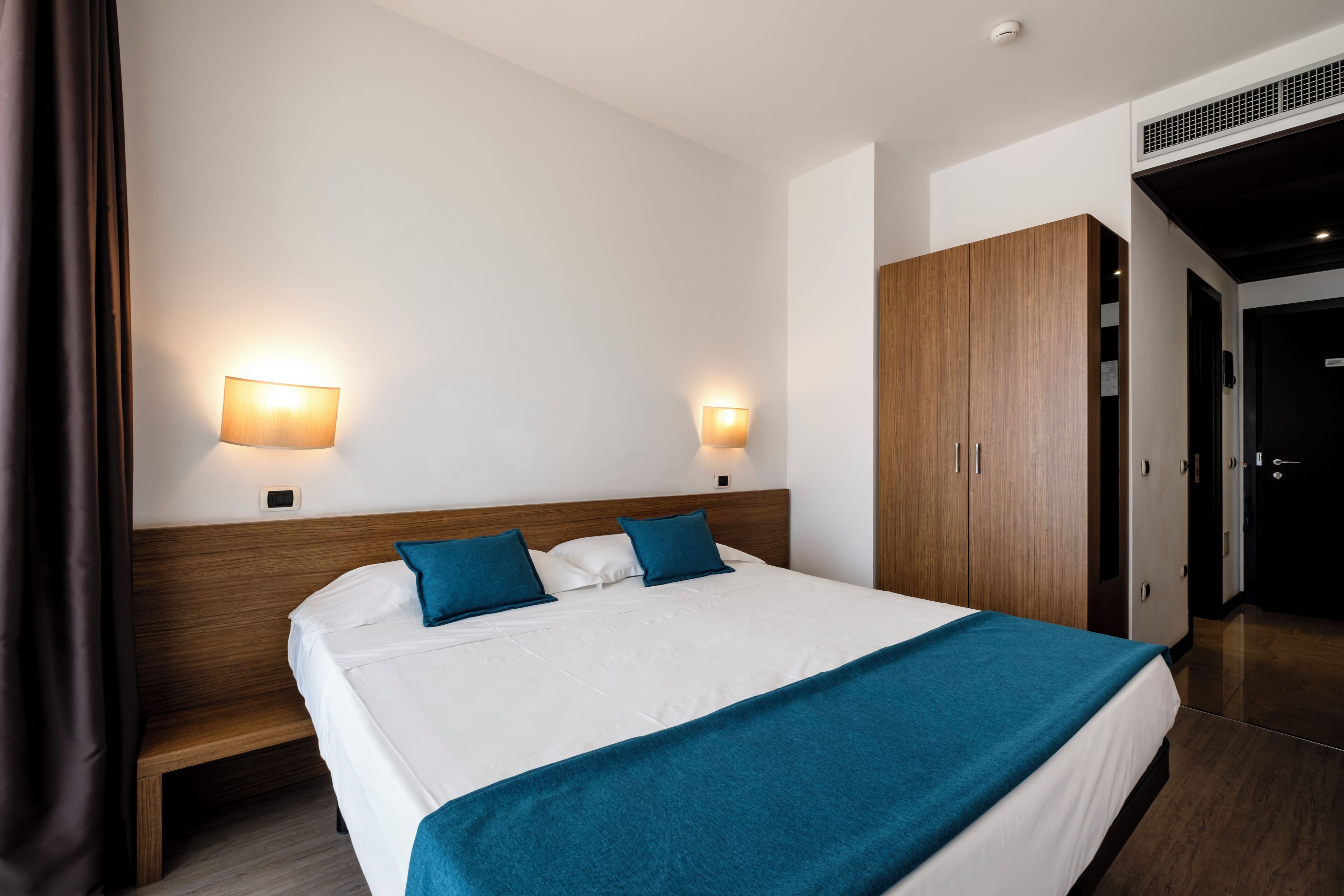 A modern hotel room with a large bed, two wall-mounted lights, a wooden wardrobe, and a bench.