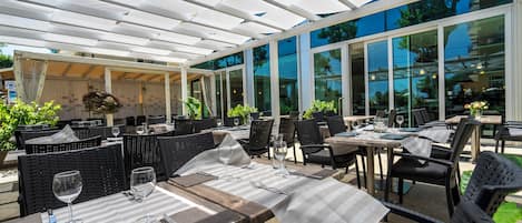 Un espacio de comedor al aire libre con mesas y sillas, ventanas amplias y una sección cubierta.