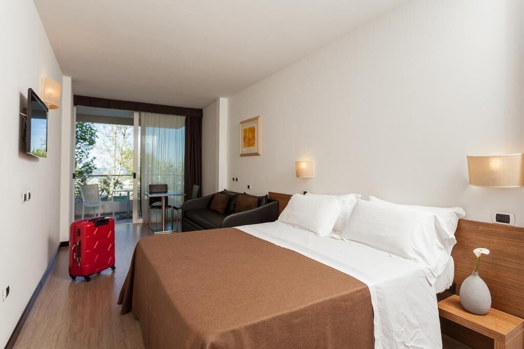 A hotel room with a bed, a desk, a chair, a red suitcase, and a view of the outdoors.