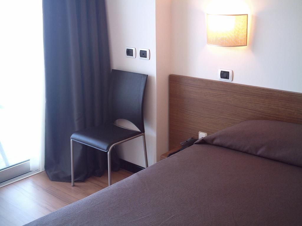A hotel room with a bed, a chair, a wall-mounted light, and a window with curtains.