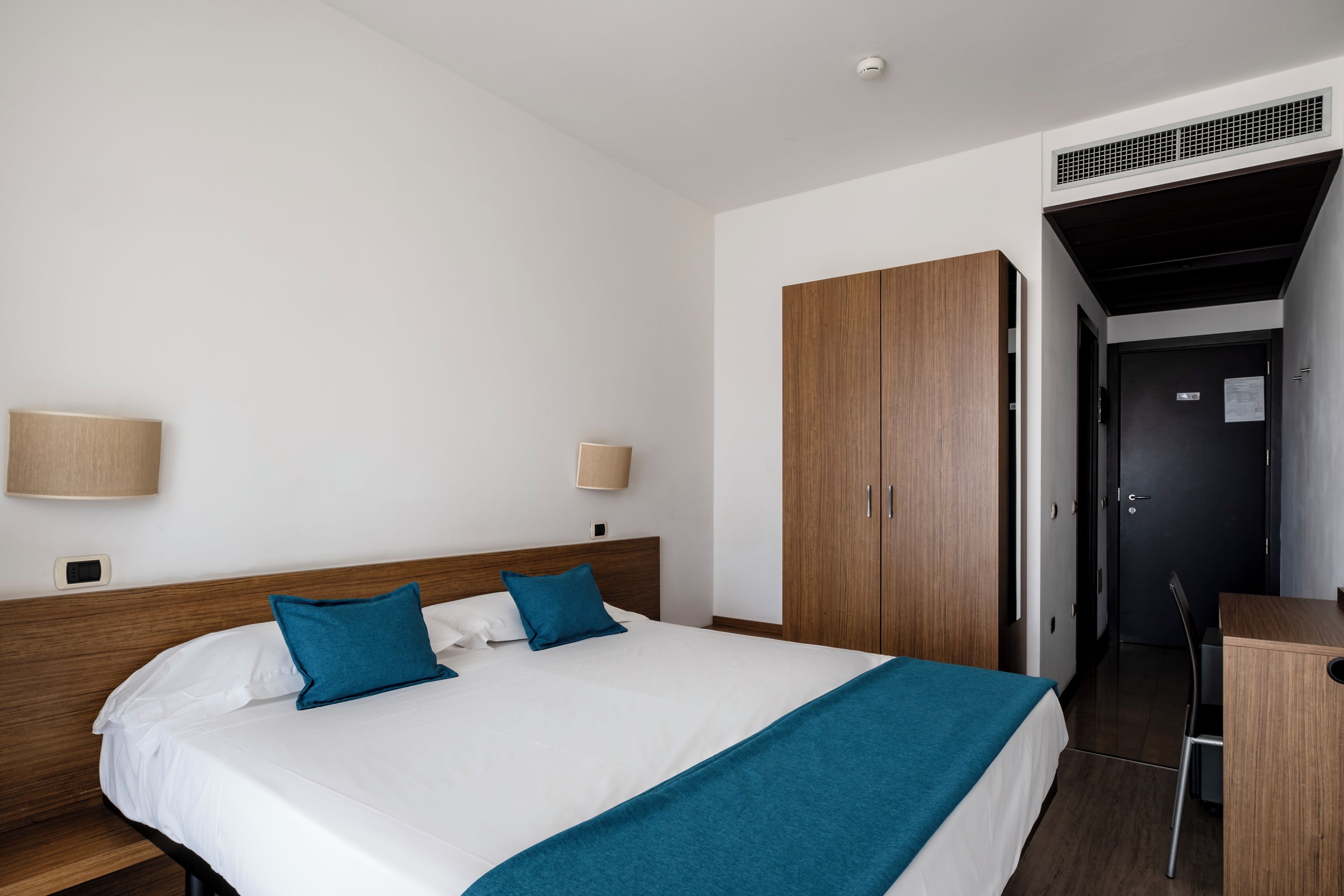 A hotel room with a bed, bedside lamps, a wardrobe, and a desk.