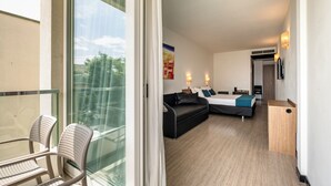 A modern hotel room with a glass balcony, a sofa bed, a desk, and a view of the outdoors.