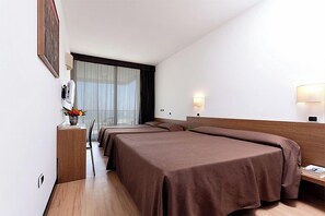 Habitació d'hotel amb dos llits, un escriptori amb cadira, una taula petita amb un ram de flors i una finestra amb cortines.