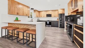 A modern kitchen with wooden cabinets, a central island with stools, stainless steel appliances, and a large window. - Relaxation+Pool & Hot Tub+Views (Comfort)