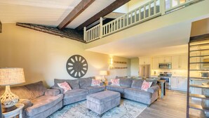 A spacious living area with a grey sectional sofa, a patterned rug, and a large wall clock. A kitchen with modern appliances is visible in the background. - Mountain Getaway Retreat - 4br + loft  (Beech Mountain)