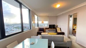 A modern living room with a glass table, grey sofa, and a TV on a wooden stand. Large windows offer a city view. - PrimeCozy & Bright 1BR Apartment (Brisbane City)