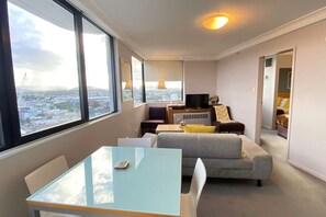 Ein modernes Wohnzimmer mit Glastisch, grauem Sofa und Fernseher auf einem Holzboden. Große Fenster bieten Blick auf die Stadt.