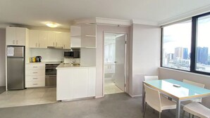 A modern kitchen with white cabinets, a stainless steel refrigerator, and a microwave. There is a small island with a sink and a coffee maker. A dining area with a table and chairs is visible, and a large window offers a view of the city skyline. - PrimeCozy & Bright 1BR Apartment (Brisbane City)