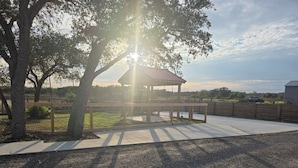 Um gazebo com cobertura metálica, rodeado por um espaço pavimentado e uma cerca.