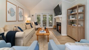 A living room with a beige sofa, a coffee table, and a fireplace. There is a television mounted above the fireplace and a bookshelf to the right.