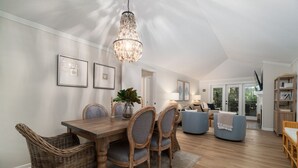 A spacious dining area with a wooden table and chairs, a hanging chandelier, and a view of the living room through an open door.