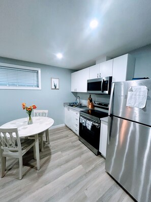 A modern kitchen with white cabinets, a stainless steel refrigerator, a microwave, and an oven. There is a dining table with chairs and a vase of flowers.