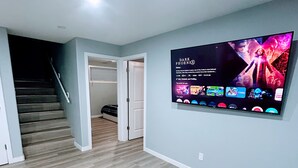 Een moderne woonkamer met een flatscreen televisie aan de muur, een grijze bank, een vloerbedekking en een trap naar een hoger niveau.