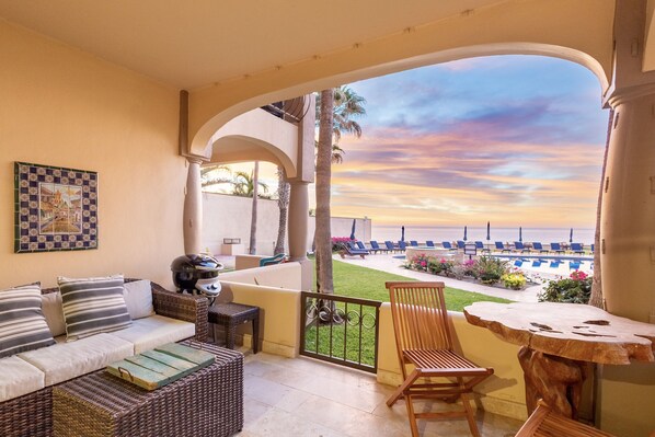 A balcony with wicker furniture, a wooden table, and a view of a pool and beach at sunset.