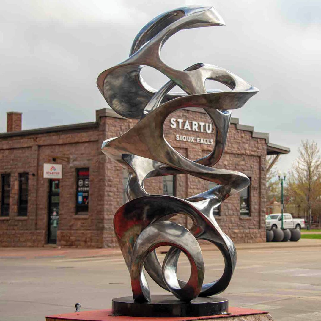 A metallic sculpture with the inscription "STARTU Sioux Falls" in front of a brick building.