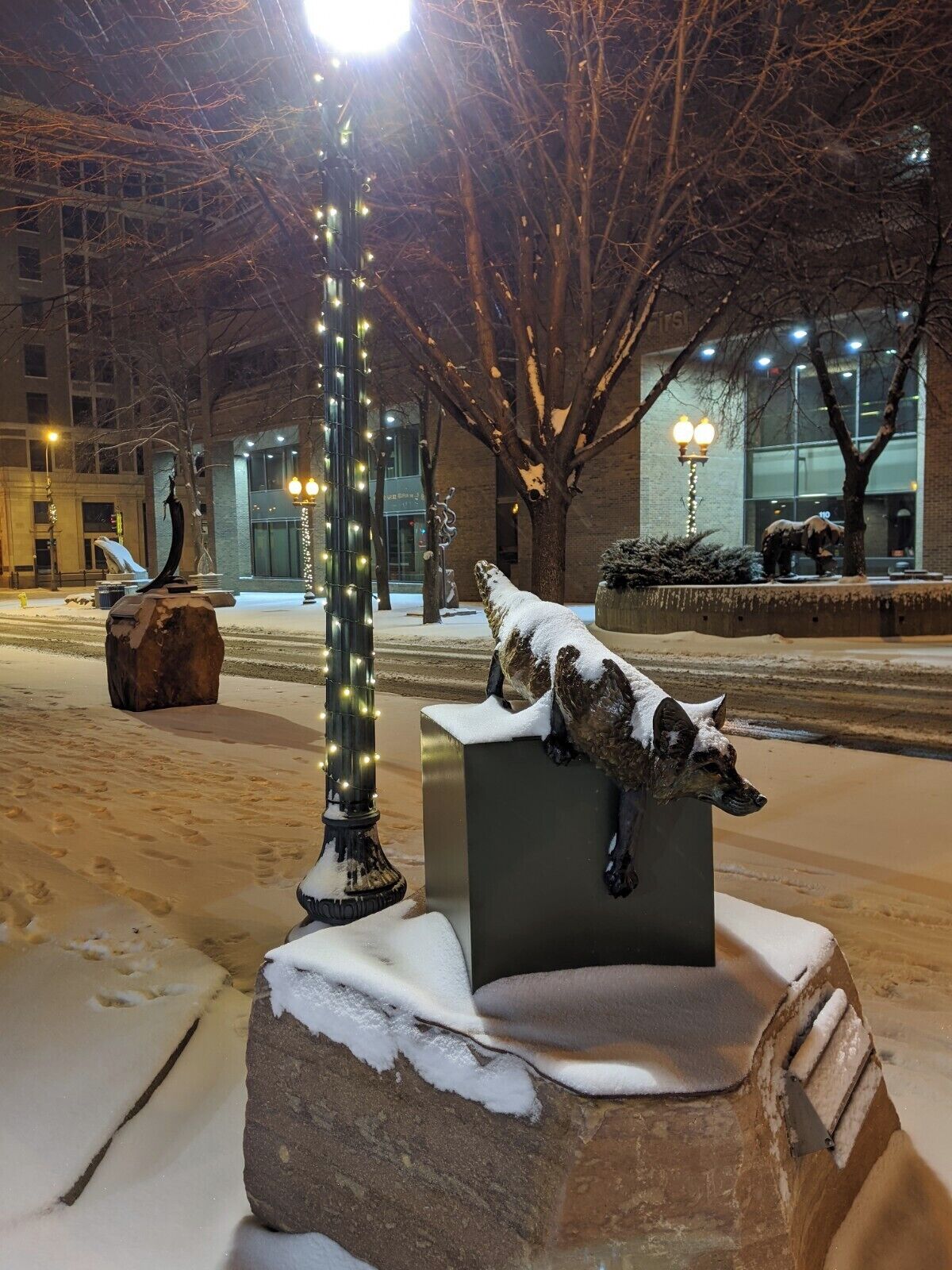 A snow-covered outdoor sculpture of a wolf on a rock, with a street lamp and bare trees in the background.