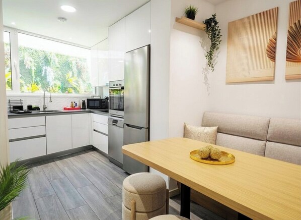 A modern kitchen with white cabinets, a stainless steel refrigerator, and a dining area with a wooden table and beige chairs.