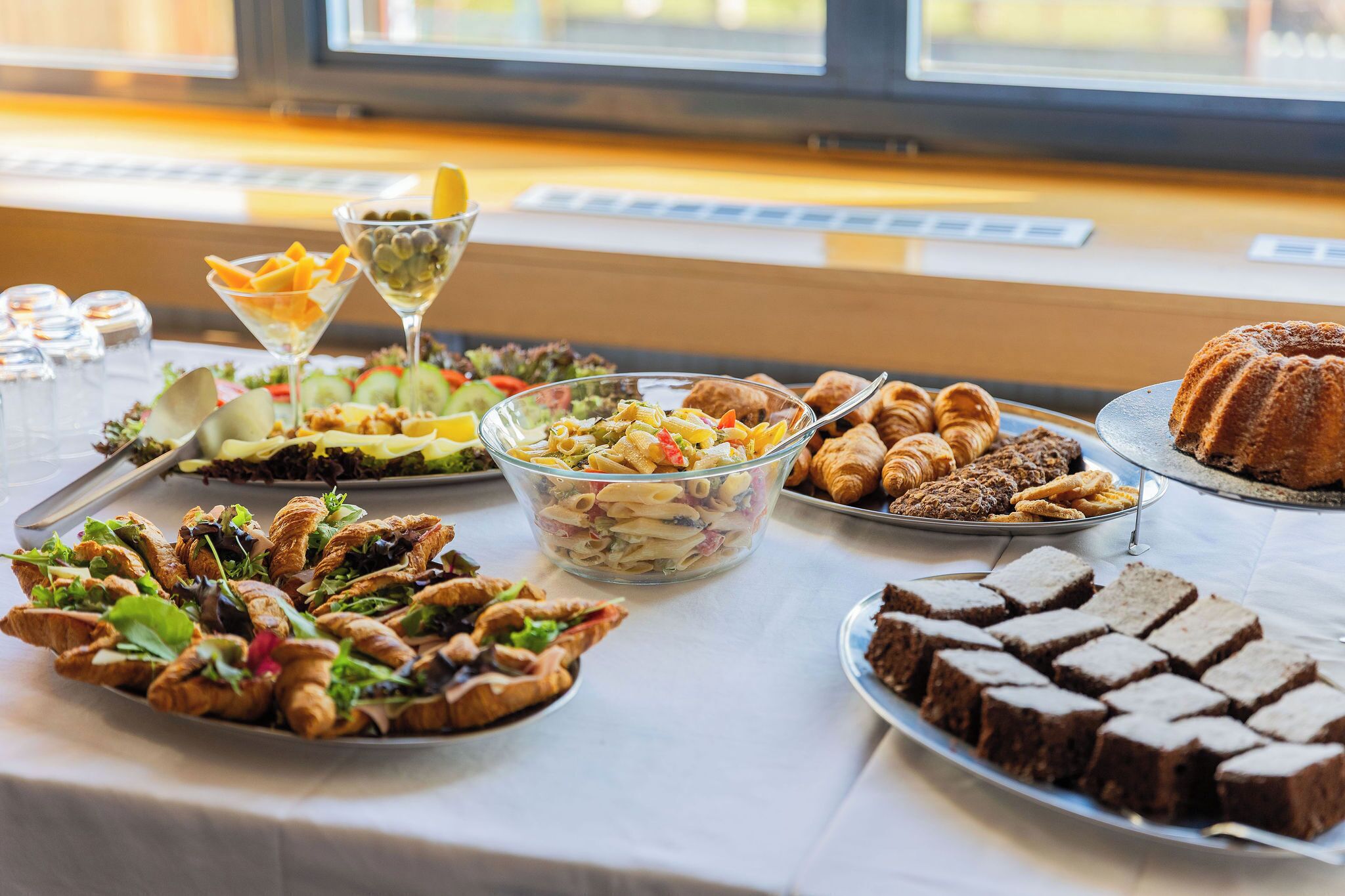 A buffet table with various dishes including pasta salad, croissants, and brownies.