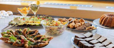 Una mesa de buffet con diversos platos, incluyendo ensalada de pasta, croissants y brownies.
