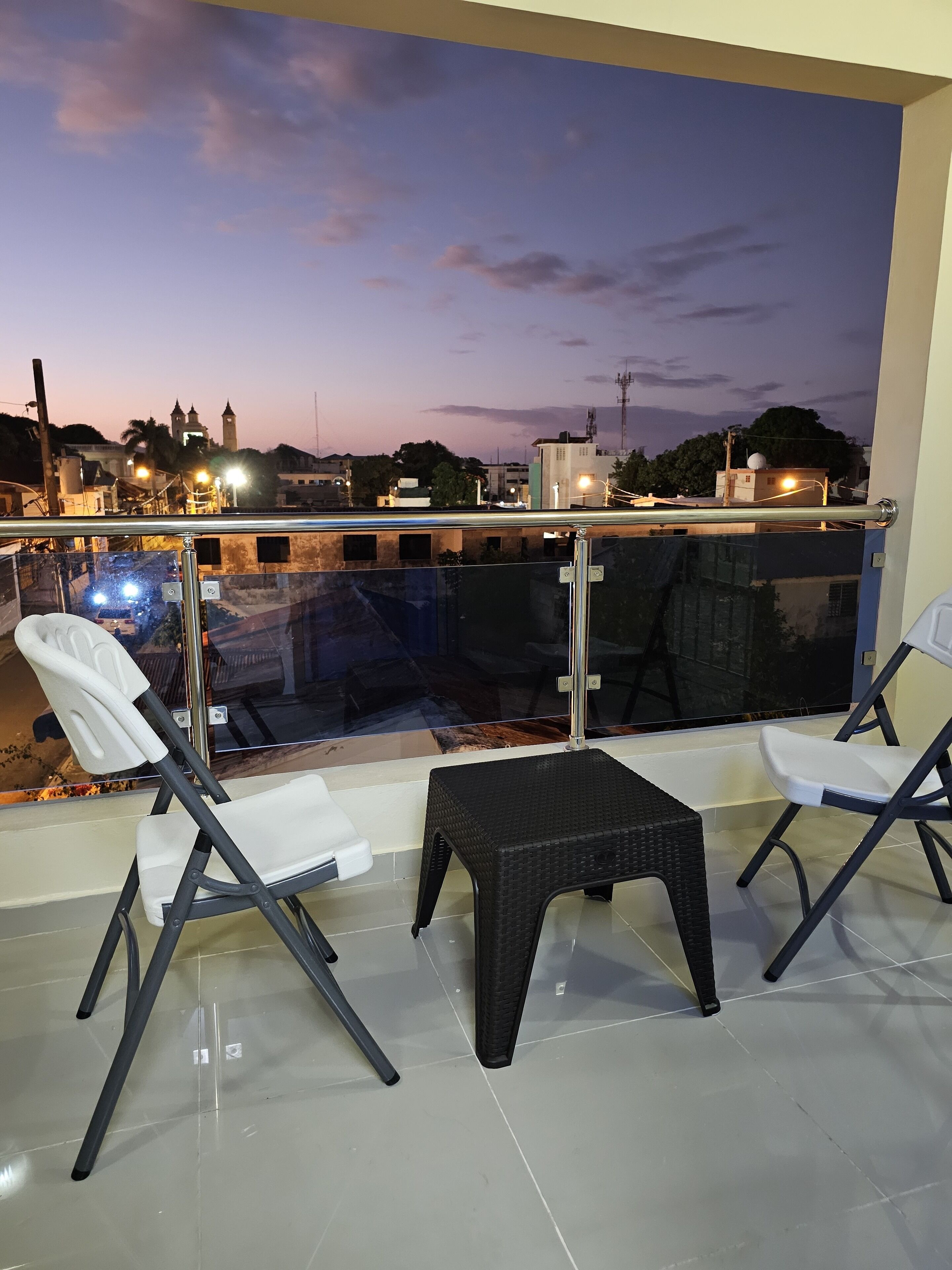 Een balkon met twee witte klapstoelen en een klein zwart krukje, met uitzicht op een stadsgezicht in de schemering, met gebouwen en een toren op de achtergrond.