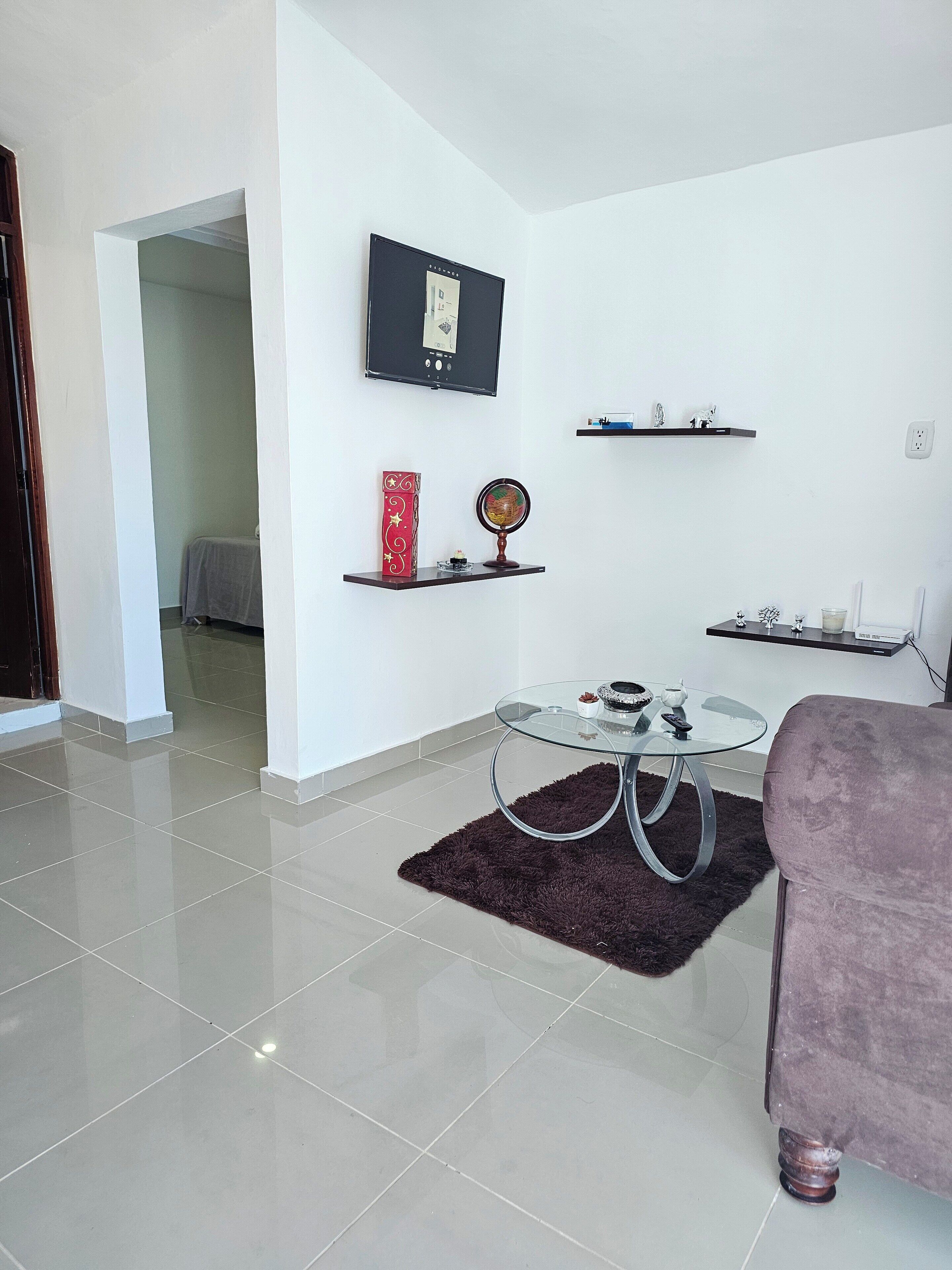A modern living room with a glass coffee table, a dark rug, a purple sofa, and a flat-screen TV mounted on the wall.