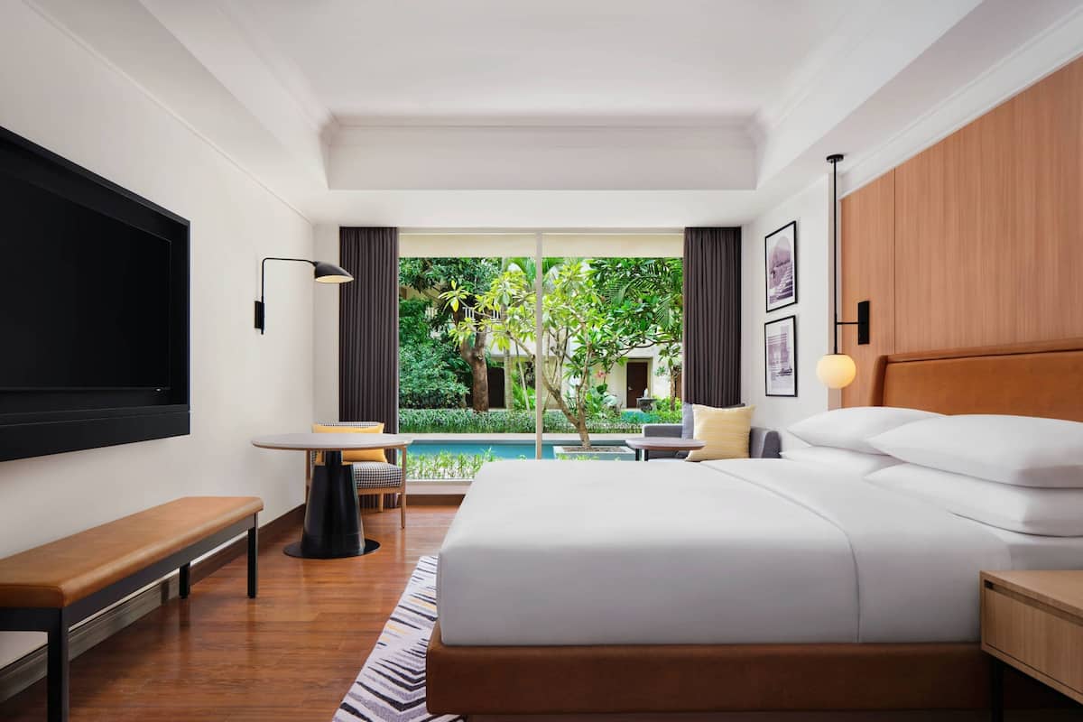 Une chambre d’hôtel moderne dotée d’un grand lit, d’un bureau, d’une banquette, d’une télévision et offrant une vue sur une piscine et un espace verdoyant.